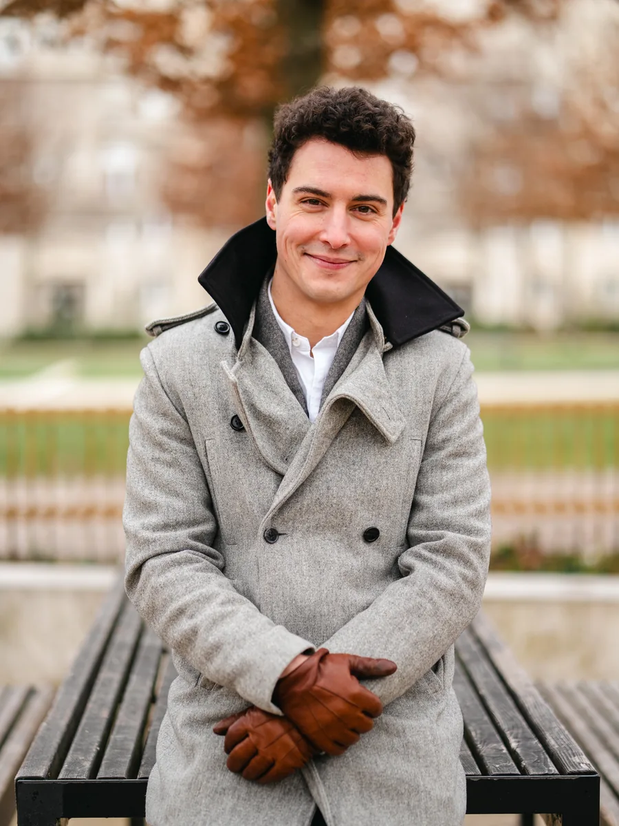 Alexis Attimont assis sur un banc, manteau gris et gants en cuir, souriant