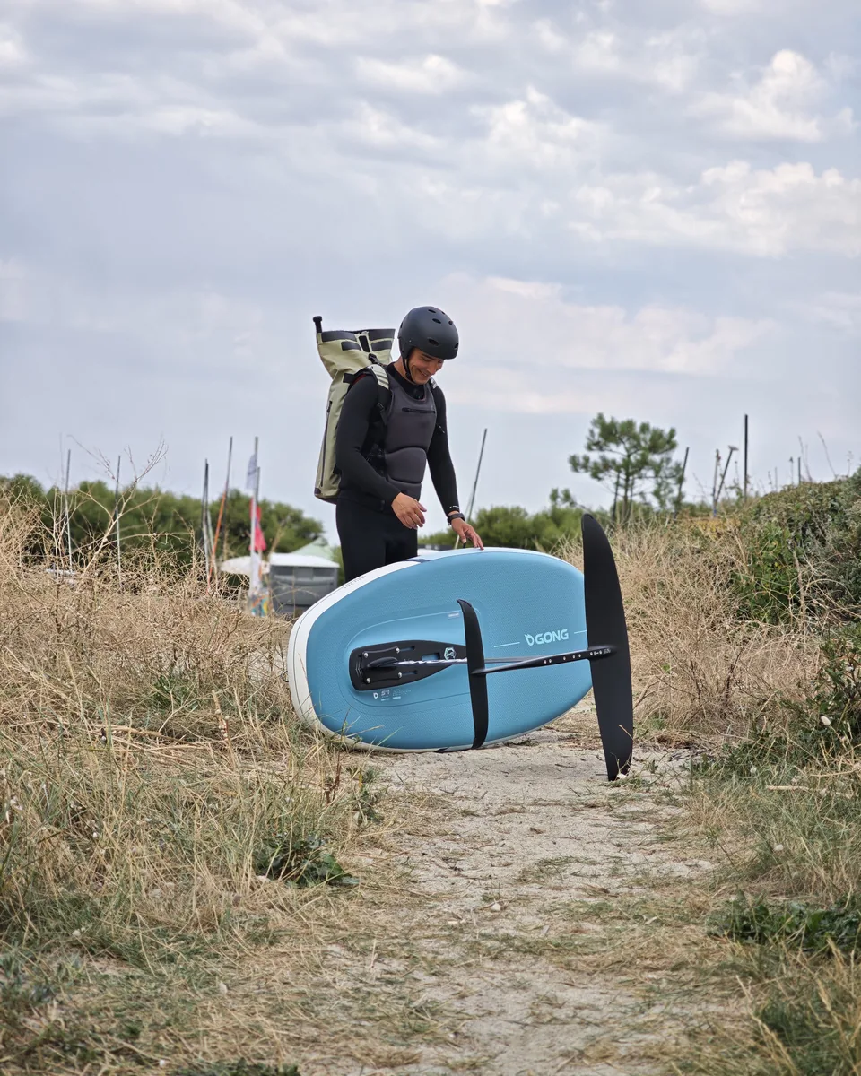 Alexis Attimont en combinaison et casque, une planche de wing foil sous le bras, sur un sentier de dune
